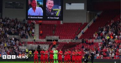 Rear view of a football team - wearing red strips - standing to attention in front of a bank of  stadium seats, with other players to the side. A screen above shows the image of two men.