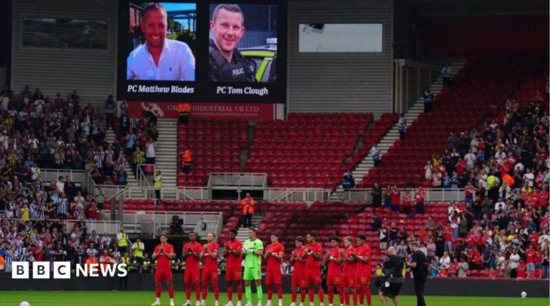 Rear view of a football team - wearing red strips - standing to attention in front of a bank of  stadium seats, with other players to the side. A screen above shows the image of two men.