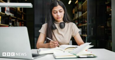 A woman sits in a university library with a laptop and two books, making notes