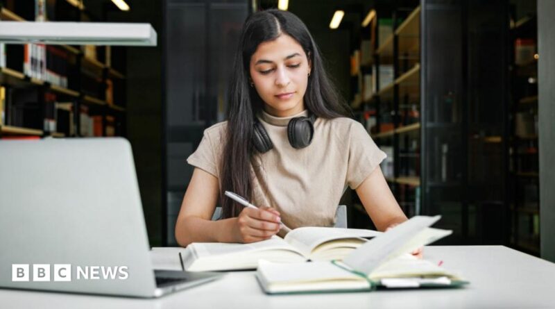 A woman sits in a university library with a laptop and two books, making notes