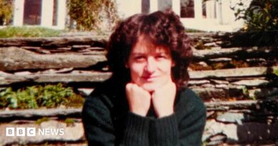 A woman sits looking at the camera with her chin resting on her balled fists. She is slightly smiling. She has curly dark brown hair and is wearing a dark green jumper. Behind her is a stone wall with some green plants growing out of it and behind that is a white house.