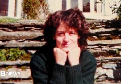 A woman sits looking at the camera with her chin resting on her balled fists. She is slightly smiling. She has curly dark brown hair and is wearing a dark green jumper. Behind her is a stone wall with some green plants growing out of it and behind that is a white house.