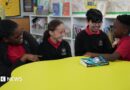 Four primary-age pupils sit around a large circular yellow table in a school library or reading area. The pupils are wearing red polo shirts with dark cardigans or jumpers. 