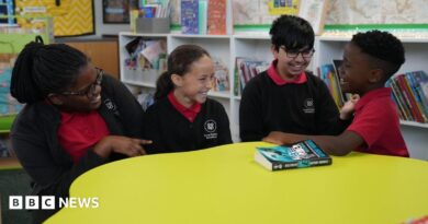 Four primary-age pupils sit around a large circular yellow table in a school library or reading area. The pupils are wearing red polo shirts with dark cardigans or jumpers.