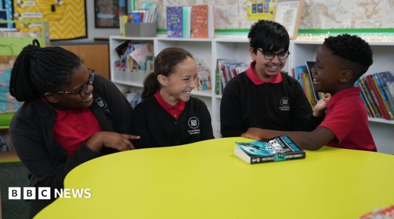 Four primary-age pupils sit around a large circular yellow table in a school library or reading area. The pupils are wearing red polo shirts with dark cardigans or jumpers.