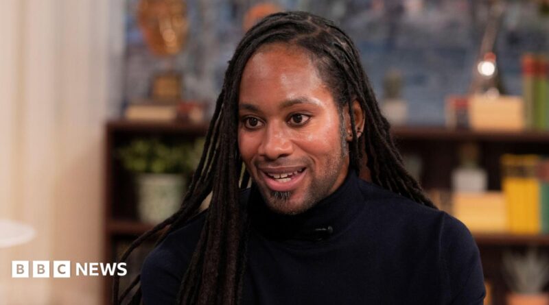 Jason Arday pictured during a TV interview in March 2023. He wears a black turtle-neck top.