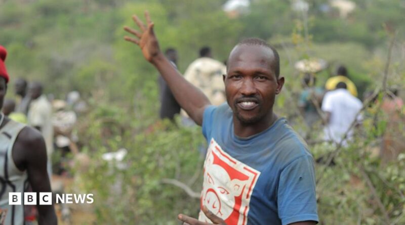 Manasse Lomachar in a blue T-shirt with a cartoon pig printed on it looks at the camera but gestures behind him with his right hand. In the background, out of focus, people can be seen working among the shrubs.
