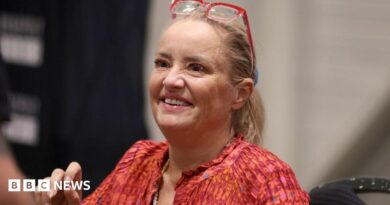 Lucy Davis, a fair-haired woman in a red top and with red-framed glasses worn on top of her head, is seen sitting down and smiling at the For The Love of Fantasy & Sci-Fi convention at Excel London on Saturday 22 August 2026