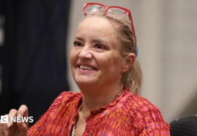 Lucy Davis, a fair-haired woman in a red top and with red-framed glasses worn on top of her head, is seen sitting down and smiling at the For The Love of Fantasy & Sci-Fi convention at Excel London on Saturday 22 August 2026