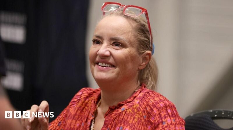 Lucy Davis, a fair-haired woman in a red top and with red-framed glasses worn on top of her head, is seen sitting down and smiling at the For The Love of Fantasy & Sci-Fi convention at Excel London on Saturday 22 August 2026