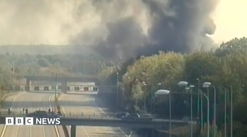 A large plume of smoke by the M25 motorway, with cars on the road across a bridge. Trees are to the left and all around the road.