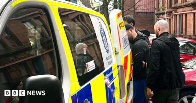 A man is escorted into the back of a blue, white and yellow Police Scotland van by two plain-clothes police officers on a residential street. Cars are parked nearby.
