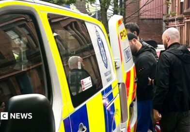 A man is escorted into the back of a blue, white and yellow Police Scotland van by two plain-clothes police officers on a residential street. Cars are parked nearby.