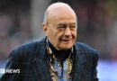 Al Fayed is wearing a suit jacket as he walks across a football pitch