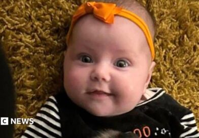 Dahlia-Rose Gartshore, a three-month-old baby girl looking at the camera, with a slight smile. She has an orange band on her head with a bow.