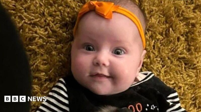 Dahlia-Rose Gartshore, a three-month-old baby girl looking at the camera, with a slight smile. She has an orange band on her head with a bow.