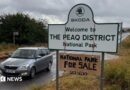 A road sign which reads SKODA WELCOME TO THE PEAQ DISTRICT NATIONAL PARK. A cardboard sign is hung around the larger sign which reads NATIONAL PARK FOR SALE CASH NEEDED.