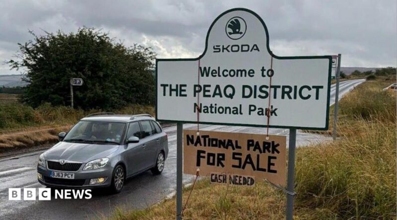 A road sign which reads SKODA WELCOME TO THE PEAQ DISTRICT NATIONAL PARK. A cardboard sign is hung around the larger sign which reads NATIONAL PARK FOR SALE CASH NEEDED.