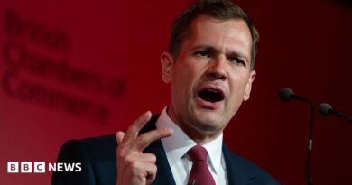 Robert Jenrick makes a hand expression as he speaks at a podium in front of a red-lit background.