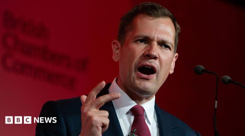 Robert Jenrick makes a hand expression as he speaks at a podium in front of a red-lit background.