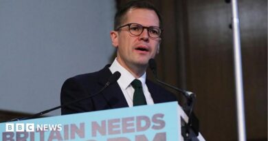 Headshot of Jenrick who speaks into two small microphones on stands on a podium in front of him that has a blue sign partially cut out of the frame, but you can read it says 'Britain needs reform'.