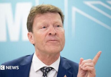 Richard Tice, in blue jacket, white shirt, and black and white tie, points with his finger while talking to reporters     