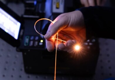 A hand holds a single strand of fiber in a darkened room, with light pouring from its tip.
