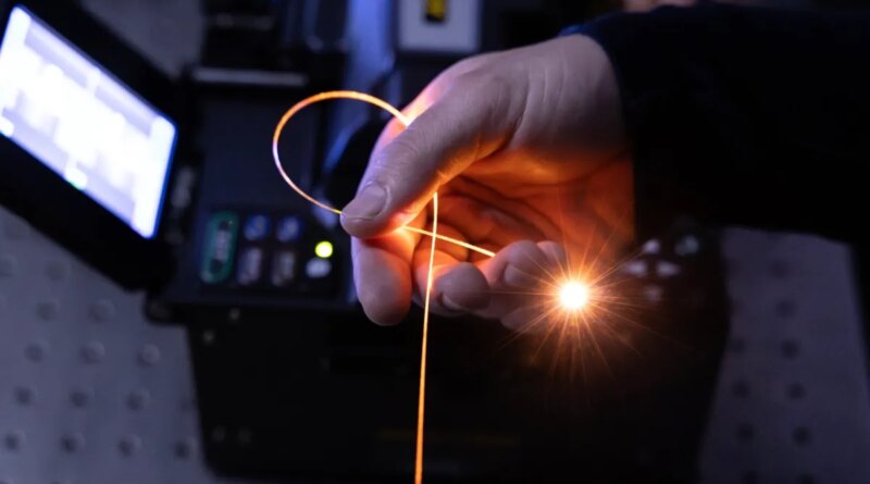 A hand holds a single strand of fiber in a darkened room, with light pouring from its tip.