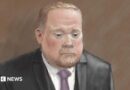 Courtroom sketch of Robert Bush in a dark three-piece suit standing alone against a plain brown background. He wears a light-coloured shirt and a purple tie. The illustration is rendered in soft pencil and pastel tones.
