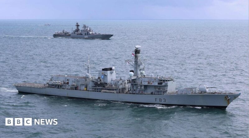 A grey Royal Navy warship is in the foreground with a grey Russian vessel in water in the background