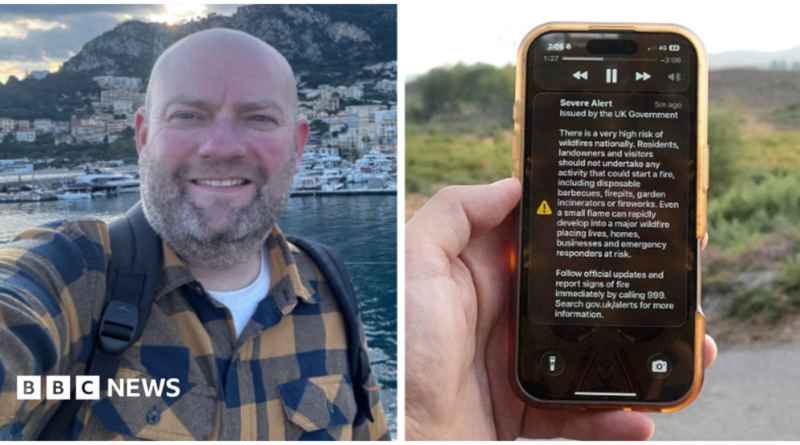A man holds a phone in front of a green field. The phone is displaying the UK government wildfire alert.