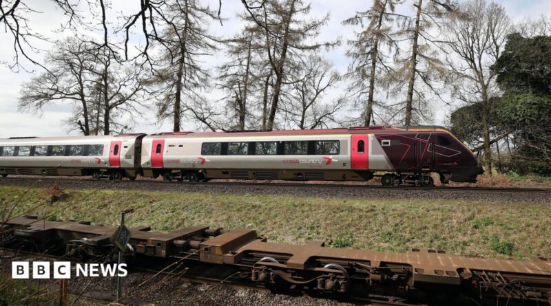 The cab and first carriage of a CrossCountry train travels on tracks in the countryside. In the background trees and fields can be seen.