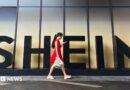 A woman in a red dress looks at her smartphone while walking past a Shein sign outside the company's office in Guangzhou, China. 