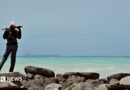 A man - with his back to the camera - stands on some rocks to the left of the picture. Holding a gun across his shoulders, he is looking out to sea where a tanker is in the far distance. 