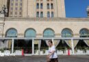 A woman wearing a red jumper and sunglasses, as well as a little bag on her right shoulder, talks on the phone in front of an exterior view of the Central Bank of the Russian Federation building on top of which the Russian flag can be seen