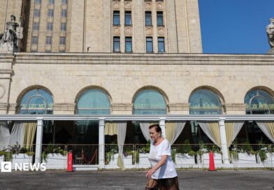 A woman wearing a red jumper and sunglasses, as well as a little bag on her right shoulder, talks on the phone in front of an exterior view of the Central Bank of the Russian Federation building on top of which the Russian flag can be seen