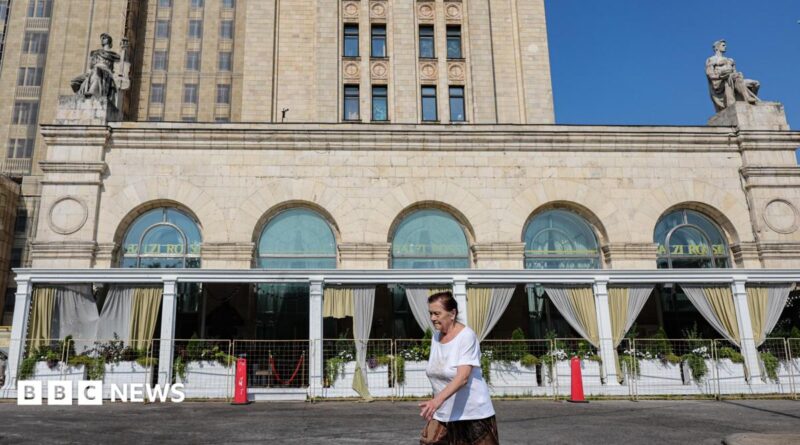 A woman wearing a red jumper and sunglasses, as well as a little bag on her right shoulder, talks on the phone in front of an exterior view of the Central Bank of the Russian Federation building on top of which the Russian flag can be seen