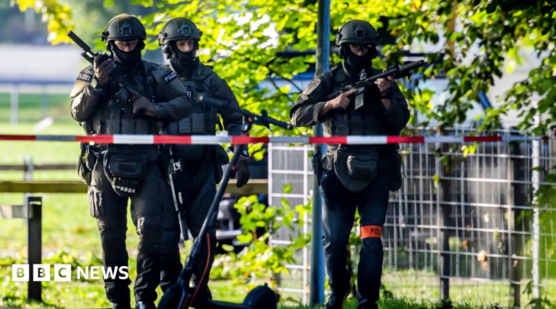 Three police officers in riot gear (helmet, bulletproof vest, knee and elbow protection) carrying guns patrol on the street, a police cordon is visible in the foreground