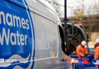 A shot of a Thames Water van. In the background there are two men wearing hi-vis clothing and hard hats and standing in an area that is boarded off by blue fences. 