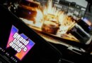 In this photo illustration, a phone displaying the Grant Theft Auto VI logo and new release date is shown in front of a screen displaying the second official trailer for the game.