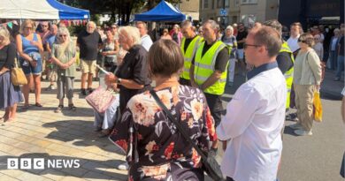 A vicar with lots of people with heads bowed in a town square daytime 