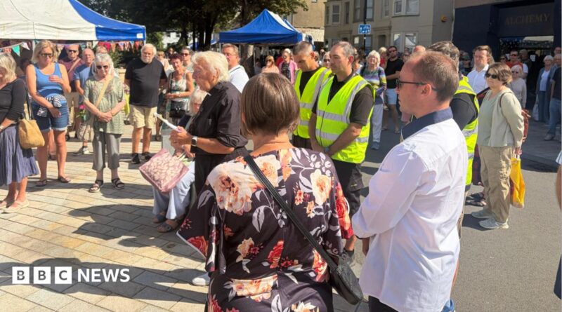 A vicar with lots of people with heads bowed in a town square daytime