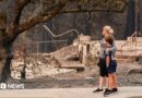 A woman dressed in black capris, grey jumper and black trousers wraps her arm around a young boy in black shorts, black top and black trainers, as both survey the damage of a burnt tree and rubble of a building