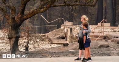 A woman dressed in black capris, grey jumper and black trousers wraps her arm around a young boy in black shorts, black top and black trainers, as both survey the damage of a burnt tree and rubble of a building