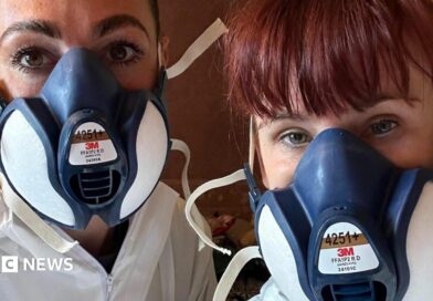 Two women in a selfie. The woman on the right has ginger hair tied back and a fringe. Her colleague has brown hair which has been tied back. They are wearing white hazmats suit and navy face masks.