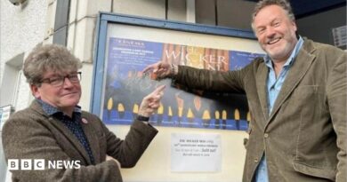 Two men in their 50s either side of a poster for The Wicker Man outside a cinema. Dominic, on the left, is in a tweed suit and has glasses and thick hair. Justin (right) is in a cord suit and has a beard. They are smiling and pointing at the poster.
