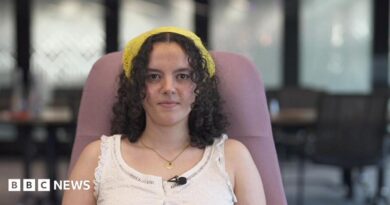Close up head and shoulders of a young woman in a sleeveless shirt looking serious. There are out of focus tables and chairs in the background.