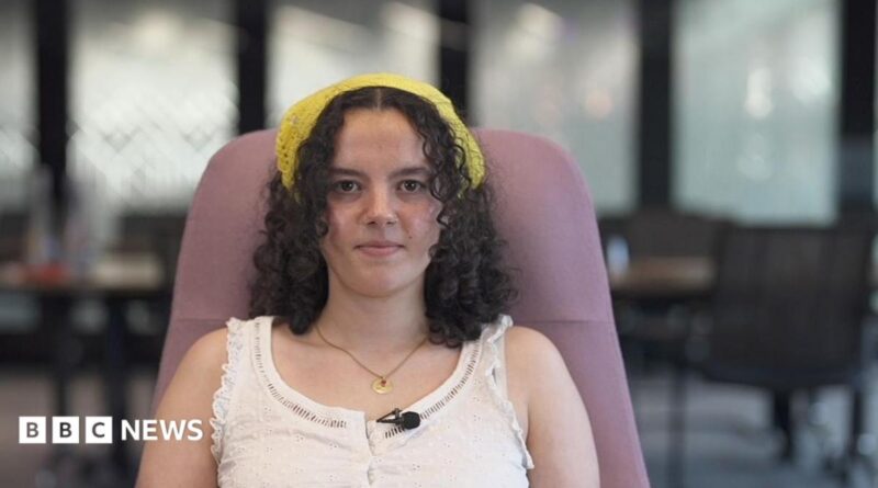 Close up head and shoulders of a young woman in a sleeveless shirt looking serious. There are out of focus tables and chairs in the background.