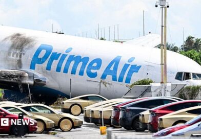 A parking lot filled with Tesla Cybercabs is seen in the foreground as the nose of a Prime Air cargo plane rests on the ground after the aircraft overran the runway and struck cars the previous day at Miami International Airport in Miami, Florida, on September 7, 2026.