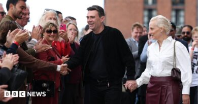 British Prime Minister Andy Burnham dressed in all black and shaking hands with a supporter in a crowed, while holding his other hand with his wife Marie-France van Heel, who is dressed in a white shirt and burgundy leather skirt.